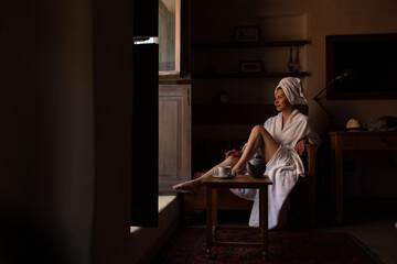 Woman in dark room sitting in robe by window. Attractive young girl in bathrobe and with towel on...