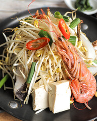 A black wok containing stir-fried noodles mixed with shrimp, tofu, vegetables, and herbs, garnished...
