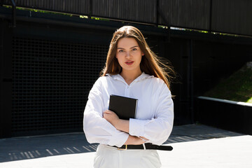 A woman in a park in a white shirt and white pants sits on a bench holding a notepad