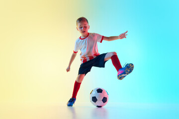 Energetic, dynamic hot of young boy in soccer uniform, kicking soccer ball mid-air in neon light against gradient background. Concept of professional sport, championship, youth league, hobby. Ad