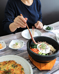 A bowl of spicy Korean tofu soup with vegetables, served alongside a bowl of white rice and a wooden spoon, placed on a rustic gray tabletop.