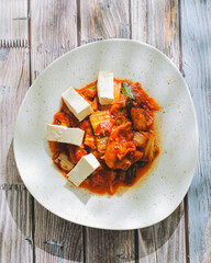 A plate of spicy Korean tofu stir-fry with vegetables sits next to a pair of chopsticks on a textured gray surface. The dish is very lively with its reddish sauce and chunky ingredients.