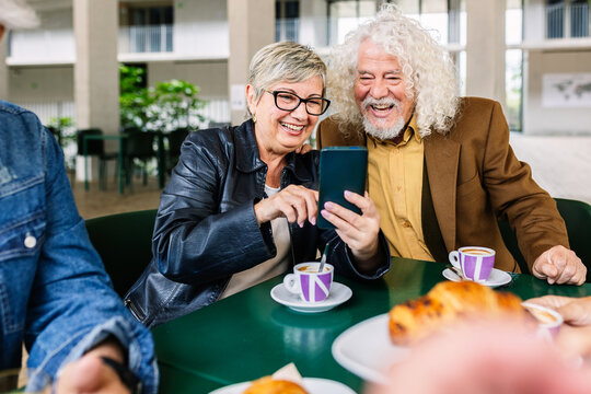 Happy senior couple of pensioners using mobile phone at cafeteria bar. Two mature people watching social media on smartphone device while enjoying coffee at restaurant.