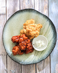 A square plate contains Korean food featuring fried chicken: two pieces plain, two pieces coated in a red sauce, served with small bowls of sauce and pickled radish, sprinkled with sesame seeds on