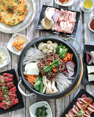 A korean food spread on a wooden table, featuring a central hot pot with noodles, vegetables, and slices of meat, surrounded by side dishes including dumplings, kimchi, and raw meats ready
