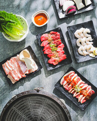Top-down view of a Korean food barbecue setup with plates of raw beef, pork, shrimp, and vegetables...
