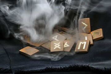 Thick gray smoke descends on wooden runes on a dark background close-up	
