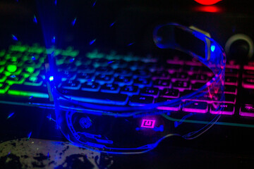 Glowing blue futuristic glasses with transparent glass lie on a multi-colored keyboard close-up	
