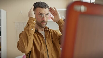 Handsome young hispanic man adjusting hair and glasses in mirror of a well-lit indoor dressing room