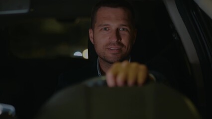 Happy young guy in suit wearing wireless earphones driving car in the evening and smiling to camera. Male taxi driver in auto. Man traveling in vehicle at night. Close up shot