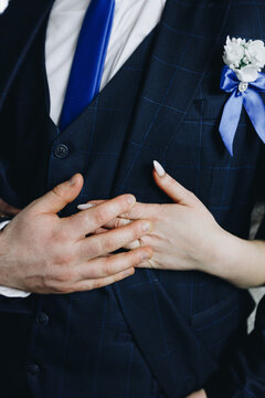 boutonniere with a white flower and a blue ribbon on a dark jacket and grrom and bride's hands