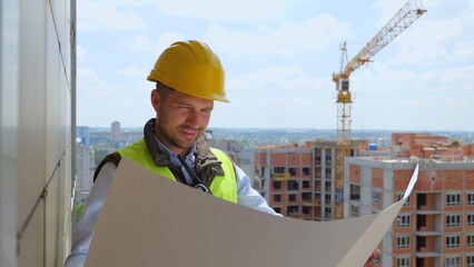 Close up of handsome young professional male constructor in helmet looking at plan draft at high...