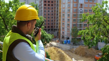 Caucasian young man construction inspector in yellow hard hat talks in walkie-talkie coordinating...