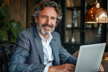Confident 50-year-old businessman works on laptop at wooden desk in modern office with warm lighting