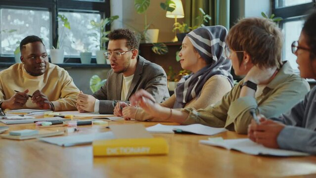 Young multi-ethnic immigrant students sitting around table and having discussion during lesson in classroom of language school