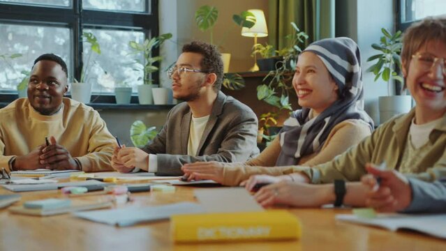 Group of joyous diverse students sitting around table in classroom, chatting and laughing on lesson in school for immigrant