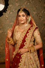 Stunning Indian bride dressed in traditional bridal lehenga with heavy gold jewellery and veil smiles tenderly in studio lighting. Wedding fashion and lifestyle.