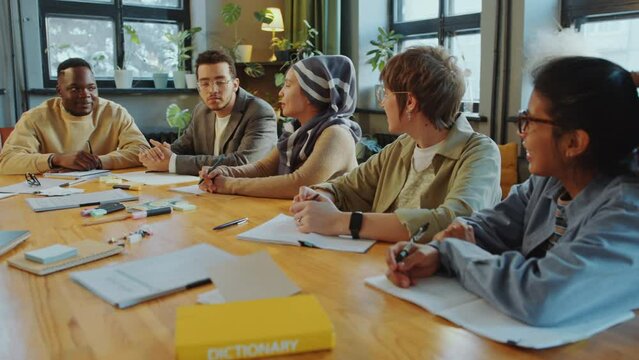 Young cheerful diverse students sitting around table in classroom and chatting during foreign language lesson in school for immigrants