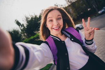Photo of pretty good mood girl wear school uniform rucksack showing v-sign tacking selfie outside...