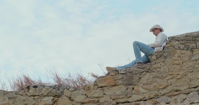 A young adult man sits on a medieval wild stone wall against a blue sky and uses a smartphone