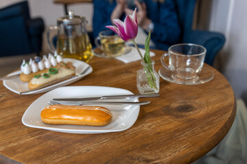 setting with a wooden table featuring an eclair on a white plate, accompanied by silver cutlery and...