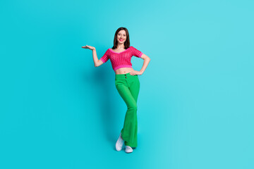 Full size photo of gorgeous nice girl wear pink top green trousers palm presenting offer empty space isolated on blue color background