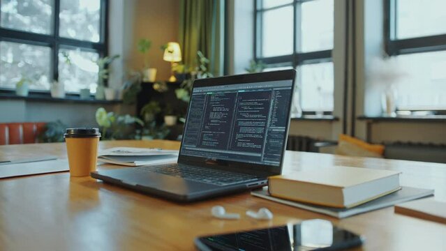 Laptop with programming code running on screen at desk with mobile phone, textbooks and disposable coffee cup in library or office. Zoom close-up shot, no people