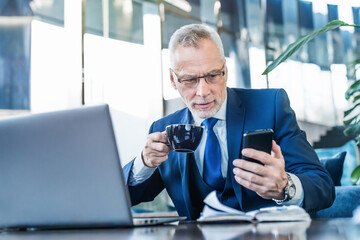 Senior male executive drinking coffee and holding smartphone while working online in office lounge....