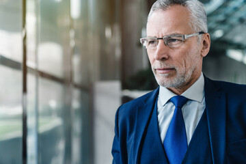 Close up portrait of senior businessman in suit standing in office lobby and looking away through...