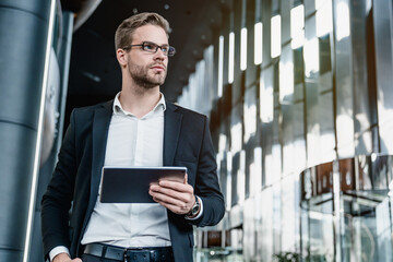 Low angle view of confident young man ceo investor wearing suit working on digital tablet while standing in business center. Businessman looking away thinking working on tech device.