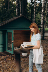 Smiling red-haired teenage girl chooses vintage books in a bookcase for bookcrossing in a city park