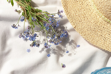 White organic cotton blanket with forget me not flowers, glass of water and straw hat. Summer picnic concept composition. 