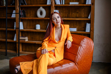 Red-haired girl in an orange business suit sitting on a leather sofa in the library