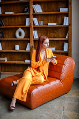 Obraz premium Red-haired girl talking on a mobile phone in an orange business suit while sitting in the library