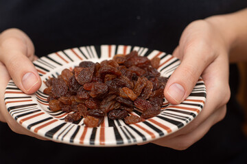 Close-up shot of a person's hands holding a small striped plate filled with juicy, dark raisins.