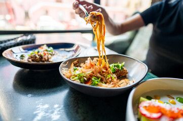 A person lifts noodles with fork from a bowl, with another dish in the background on a table. The focus on the action of eating makes it relevant for culinary themes
