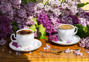 A bouquet of purple lilac flowers and two white vintage coffee cups with spoons on a wooden background. Spring still life in sunlight.