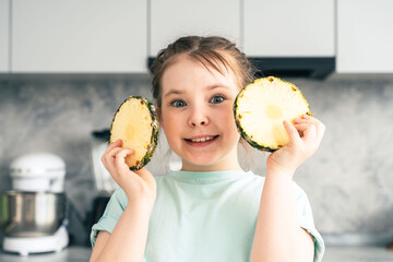 Cheerful little girl with pineapple cut into circles standing in the kitchen at home. She looks...