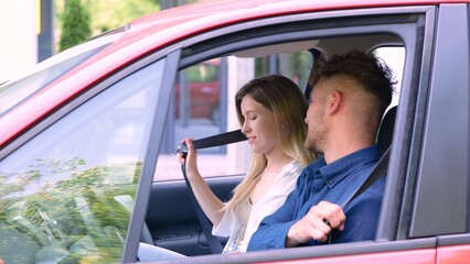 Camera approaching young male and female couple sitting inside the vehicle and fastening seat belts. Positive pretty woman in car with young beloved man. Love relations. Travel concept