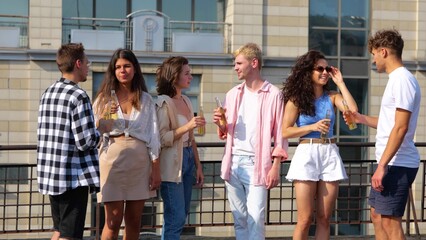 Group of young casual people standing outdoor with drinks at rooftop party on sunny day speaking...