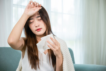 Sick woman sitting under blanket on sofa and sneeze with tissue paper in living room, Asian young...