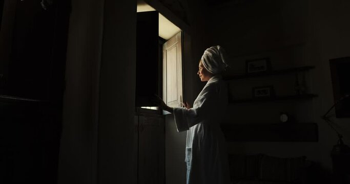 Woman in dark room old hotel standing in robe. Young leady in bathrobe and with towel on her head, looking in window and smiling. Girl has opened the shutters and is standing looking out window