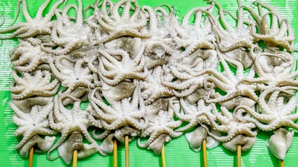 Octopus skewers are carefully arranged on bright green cutting board at street food stall. Fresh,...