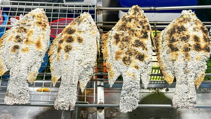 Salt-crusted grilled fish rest on grill rack at street food market. Their crispy, charred skin...