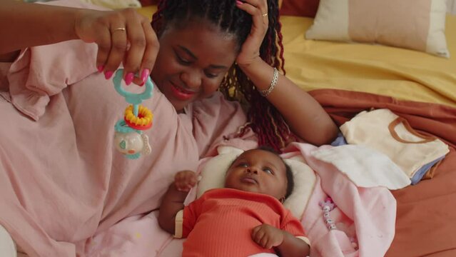 Medium shot of black mother shaking rattle over babys head lying together on bed at home