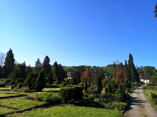 Schotterweg mit Gräbern, Bäumen und Friedhofsmauer vor blauem Himmel im Sonnenschein auf dem...