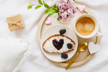 Eine Tasse Kaffee, eine Scheibe Brot mit Konfitüre in Herzform und Blumen auf einem Holz Serviertablett auf einer weißen Bettdecke. Draufsicht, süsses Frühstück.