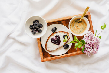 Eine Kaffeetasse, eine Scheibe Brot mit Konfitüre in Herzform und frische Blumen auf einem braunen Holz Serviertablett. Weiße Bettdecke, Draufsicht, süsses Frühstück.