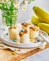 Slice banana pieces with peanut butter, colorful sprinkles, and a knife on a light wooden table. Space for text. Creative idea for kid breakfast, healthy snack for kids.