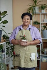 Vertical portrait of kind Black senior woman holding green plant and smiling at camera while enjoying gardening